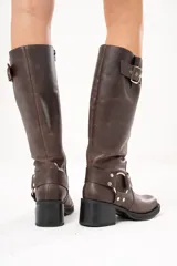 Bota biker de caña alta color marrón, confeccionada en cuero. Presenta hebilla en la parte superior, detalle de aro en la parte inferior y cierre lateral.