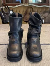 Bota corta de cuero con efecto desgastado, color marrón oscuro con matices bronceados. Presenta un diseño de caña arrugada y una tira lateral ajustable con hebilla metálica plateada. Cuenta con suela de goma gruesa y taco cuadrado.
