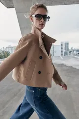 Chaqueta corta de paño en color beige, con cuello subido, manga larga y cierre frontal cruzado con botones oscuros. Presenta bolsillos delanteros de vivo.