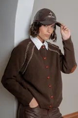 Saco de punto color beige, con cierre frontal de botones marrones y detalle de costuras decorativas oscuras en los hombros. Presenta un diseño clásico con cuello redondo.