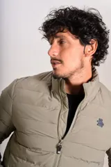 Campera puffer de corte clásico en color beige, con cierre frontal, cuello alto y puños elásticos. Presenta un diseño acolchado horizontal y un pequeño logo bordado en el pecho.