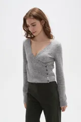 Sweater de punto gris con cuello en V, diseño cruzado y cierre lateral con botones. Presenta mangas largas y un ajuste al cuerpo con detalle acanalado en la cintura.