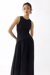 Vestido midi negro sin mangas, con cuello redondo y diseño ajustado en la parte superior de punto acanalado. Cuenta con una costura fruncida en diagonal bajo la cintura y una falda larga con vuelo de tejido fluido.