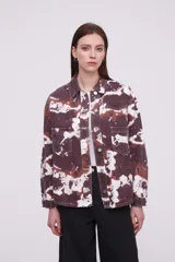 Campera tipo camisa de manga larga con cierre frontal de botones metálicos, bolsillos frontales con solapa y diseño estampado tipo tie-dye en tonos marrones y blancos.