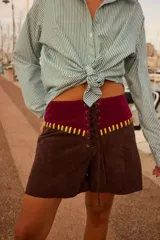 Shorts de cuero marrón con cintura alta de terciopelo burdeos, cordones frontales y apliques de cuero amarillo.
