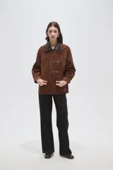 Chaqueta de gamuza color marrón con cuello camisero en cuero oscuro, cierre frontal con broches metálicos tipo gancho y bolsillos delanteros aplicados.