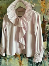 Camisa de algodón color rosa suave con textura de rayas verticales sutiles. Presenta un cuello grande con volantes que se extiende hacia el frente y mangas largas con puños abotonados.