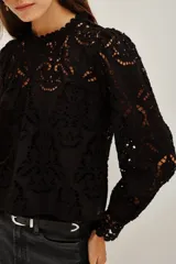 Camisa de encaje negro con cuello alto y mangas largas abullonadas. Presenta un diseño calado con motivos florales y cierre posterior.