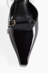 Sandalia negra de charol con punta fina, taco kitten y pulsera ajustable al tobillo. Presenta una tira delantera con hebilla dorada.