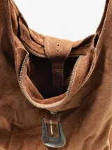 Cartera tipo hobo de diseño amplio y estructura flexible, confeccionada en gamuza color marrón con acabado desgastado. Cuenta con un asa única para llevar al hombro y un cierre frontal con detalle de hebilla metálica decorativa.