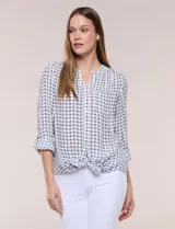 Blusa estampada marca Zac & Rachel, de color blanco con diseño geométrico en tonos oscuros. Presenta cuello en V, cierre frontal con botones, bolsillo aplicado en el pecho, mangas largas con presilla para regular el largo y detalle de nudo en el dobladillo delantero.