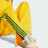 Pantalón deportivo Adidas de corte holgado, color amarillo brillante con las clásicas tres tiras verdes en los laterales y el logo del trifolio bordado en la pierna izquierda. Cuenta con cintura elástica y bolsillos laterales con cierre.