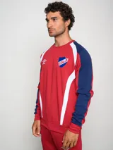 Buzo deportivo de manga larga, color principal rojo, con mangas azul marino y detalles en blanco que bajan desde el hombro hasta el puño. Presenta cuello redondo, logo Umbro y escudo del Club Nacional de Football en el pecho.