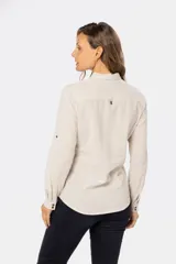 Camisa blanca de manga larga con cuello y tapeta de medio botón. Presenta dos bolsillos frontales con tapa y botones metálicos. Las mangas tienen presillas para sujetar el puño enrollado.