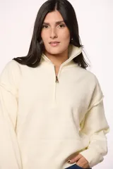 Buzo tejido de punto en color crema, con cuello alto y cierre frontal parcial. Presenta mangas largas con costuras caídas y puños acanalados.