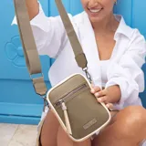 Phone bag beige tipo morral, con bolsillo frontal con cierre, ribetes negros y correa gruesa de galón regulable y desmontable.