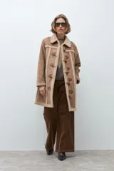Abrigo de estilo duffle coat en simil gamuza color tostado, con interior y bordes de piel sintética al tono. Presenta cierre frontal mediante alamares de cuero sintético y madera, cuello solapa y mangas largas.