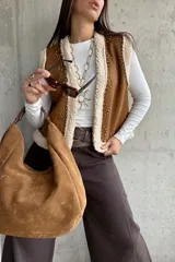 Chaleco de gamuza color camel con interior de corderito sintético, diseño sin mangas, borde frontal con ribete de corderito y detalle de tachas metálicas decorativas.