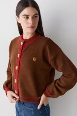 Cardigan de lana color marrón con detalles de bordes en color rojo. Presenta cierre frontal con botones, mangas largas con puños ajustados y un pequeño bordado decorativo en el pecho.