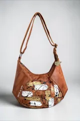 Cartera bandolera de cuero tostado con diseño de gran capacidad, líneas orgánicas y arte pintado a mano en el frente en tonos blanco, dorado y negro. Cuenta con manija regulable para usar al hombro o cruzada y detalle de llavero metálico con logo.