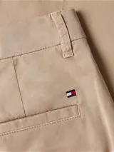 Pantalón chino color beige de sarga elástica, corte slim, con cierre de cremallera y botón, bolsillos traseros de ojal y logo de Tommy Hilfiger bordado en la parte trasera.