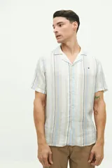 Camisa de manga corta con cuello cubano, confeccionada en una mezcla de lino y algodón con textura. Presenta un diseño a rayas verticales en tonos beige y celeste sobre fondo blanco, con cierre frontal de botones y logo bordado en el pecho.