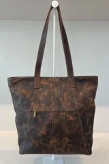 Bolso tipo shopper de cuero color suela con textura grabada, detalle de costuras laterales trenzadas y placa metálica con logo de la marca. Cuenta con doble asa larga para colgar al hombro, cierre principal y múltiples bolsillos internos y externos.