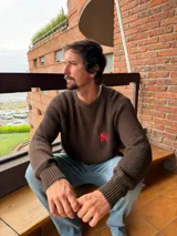 Sweater de tejido de punto acanalado en color marrón, con cuello redondo, mangas largas y bordado distintivo en color rojo en el pecho.