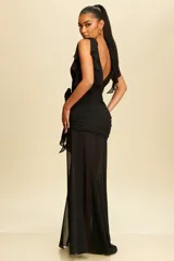 Vestido largo de fiesta color negro, confeccionado en organza con escote en V y breteles con volados. Presenta un diseño ajustado con fruncidos en la cintura, detalle de flor decorativa lateral y una falda con tajo profundo que aporta movimiento.