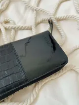 Billetera grande de cuero con cierre metálico perimetral. Presenta un diseño combinado con una sección superior de cuero liso brillante y una sección inferior de cuero con textura de reptil. Incluye el logo de la marca grabado en la parte frontal.
