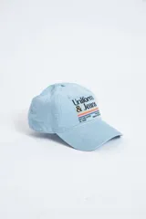 Gorra de pana celeste con visera curva y bordado frontal con el texto "Uniform & Jeans" y rayas horizontales de colores. Ajuste trasero con hebilla metálica.