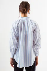 Blusa de algodón blanco con diseño de rayas verticales y textura de pequeños puntos en relieve. Presenta cuello alto con volados, cierre frontal con botones forrados y mangas largas abullonadas con puños ajustados.