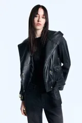 Campera de cuero ovino con capucha amplia integrada, cruce asimétrico con cierre metálico y cinturón regulable en la cintura. Presenta mangas largas con cierres metálicos en los puños y diseño estructurado.