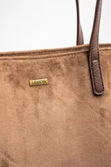 Bolso tipo tote de gran tamaño en color camel, confeccionado en cuero vegano con acabado efecto gamuza. Presenta doble asa larga para llevar al hombro y una placa metálica pequeña con el logo de la marca en el frente.
