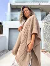 Ruana tejida en color beige, con diseño de trenzas verticales y terminación de flecos en el ruedo. Presenta una caída holgada y abierta al frente.