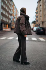 Anorak corto acolchado color marrón chocolate, con cuello subido, manga larga con puños elásticos y cierre frontal con botones ocultos y lazada lateral.