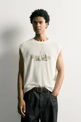 Camiseta tank blanca de corte relajado, con textura en relieve, cuello redondo y sisas amplias. Cuenta con un diseño bordado y estampado en el centro del pecho con las inscripciones "Slow Vibes" y "After Hours" en tonos verde y negro.