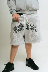 Bermuda de felpa color gris claro con efecto lavado (washed). Presenta un corte ancho con cintura elástica y cordón beige. El diseño frontal incluye un estampado serigrafiado en negro con texto y motivos florales/ornamentales en bordado blanco. Posee remaches metálicos cromados en los laterales y bolsillos.