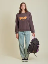 Mochila marrón con detalles en violeta. Tiene un bolsillo frontal superior con cierre, un bolsillo lateral de red y el logo de Rusty Worldwide estampado en naranja.