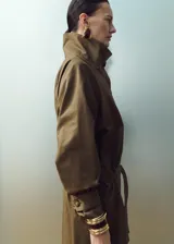 Chaqueta de gabardina color verde militar, de corte oversize con cuello alto abotonado de manera asimétrica, mangas largas con puños ajustables y cinturón ajustable del mismo tejido.