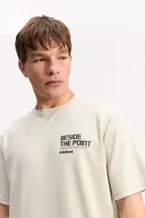 Remera de manga corta color beige, con estampa en la espalda que incluye un paisaje marino de atardecer y texto.