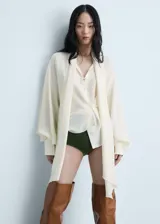 Blusa de tejido fluido en color crema, con diseño oversize, cuello en pico con lazo integrado, cierre frontal de botones y mangas largas con puños abotonados.