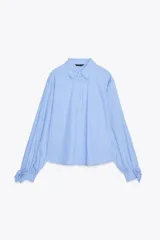 Camisa corta de manga larga, color celeste con finas rayas blancas verticales. Posee cuello solapa y puños anchos ajustados con lazos del mismo tejido.