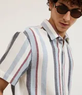 Camisa de hombre de manga corta con cuello tradicional y abotonado frontal. Presenta un tejido texturizado con un patrón de rayas verticales en tonos beige, blanco y marrón claro.