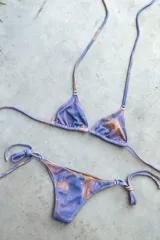 Conjunto de bikini con corpiño triangular de atar con apliques de argollitas y bombacha vedetina de atar con apliques de argollitas. El tejido presenta un estampado tie-dye o marmolado en tonos lila/azul y naranja/beige.