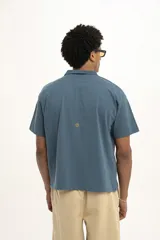 Remera de manga corta color verde oscuro, estilo polo con cuello y abertura frontal. Presenta un bordado central con el texto "PulauCrew" y un laurel en la parte superior.