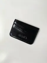 Tarjetero de cuero vacuno color negro con acabado brillante. Cuenta con cuatro ranuras para tarjetas y un bolsillo central.