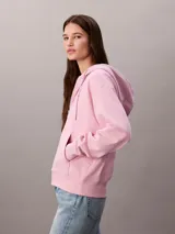 Buzo con capucha de Calvin Klein, de corte cropped y color rosado, confeccionado en mezcla de algodón y poliéster. Cuenta con cierre frontal de cremallera, cordones ajustables en la capucha y bolsillos tipo canguro. Presenta el logo de la marca bordado discretamente en el pecho.