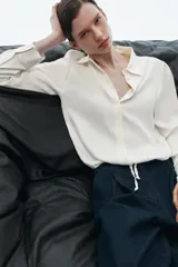 Camisa de satén con cuello solapa, manga larga y cierre frontal con botones. Presenta un diseño con bajo ajustable mediante cordones.