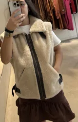 Chaleco de piel sintética en color beige, con cierre frontal y detalles en cuero marrón oscuro. Presenta cuello solapa y correas ajustables en los laterales.